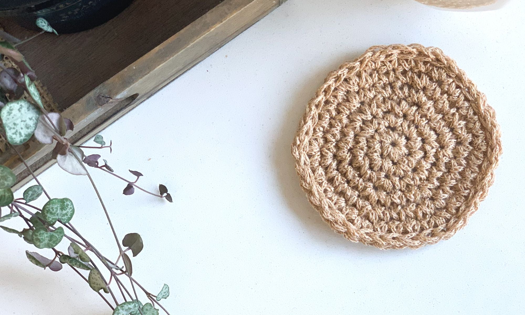 A sparkly golden coloured crochet coaster made using the magic circle spiral technique laying flat on a desktop with a trailing plant nearby and wooden tray.