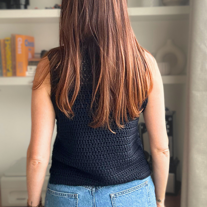 A woman viewed from behind wearing a sleeveless black crocheted top and blue jeans, standing in front of white shelves in an indoor setting.