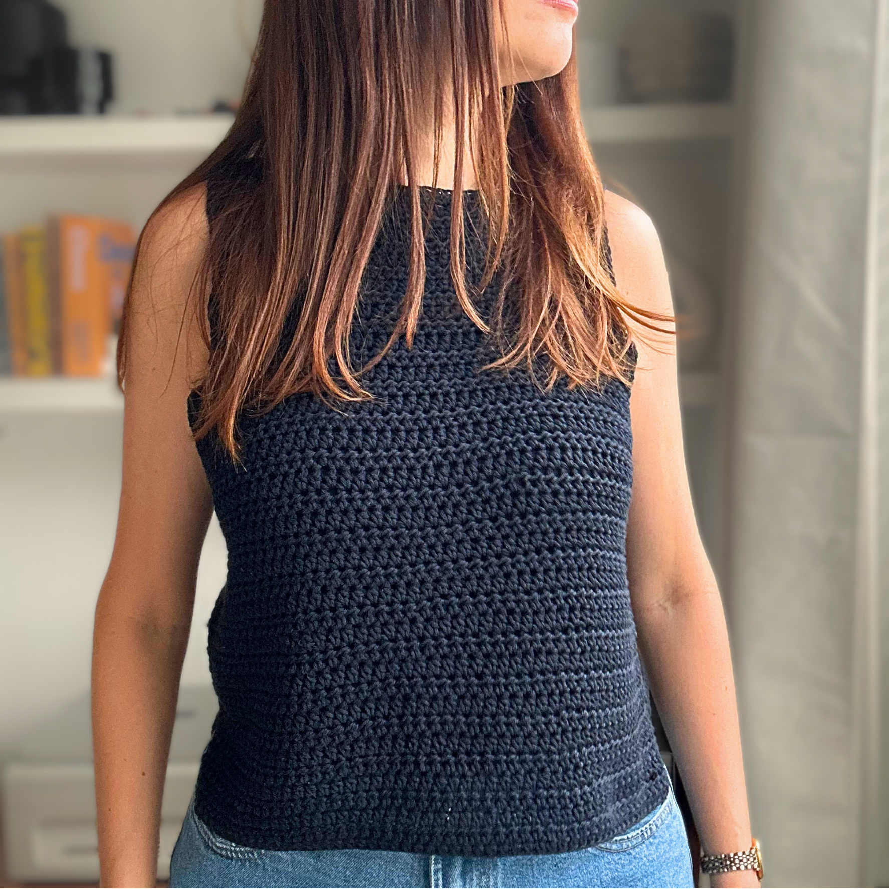 A woman wearing a sleeveless, dark navy blue crocheted top, shown from a side-back angle in an indoor setting with bookshelves and window blinds in the background.